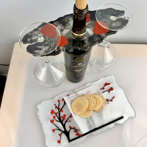 May include: A wine serving set with a bottle of wine, two wine glasses, and a serving tray. The tray has a black tree design with red heart-shaped accents and a stack of crackers.