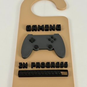 May include: A beige door hanger with the words "GAMING IN PROGRESS" in black block letters. A gray video game controller is centered on the hanger. A black loading bar is below the text.