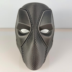May include: A black Deadpool mask with a textured surface and white eye openings. The mask has a detailed design with angular features and a distinct shape, likely for cosplay or display.