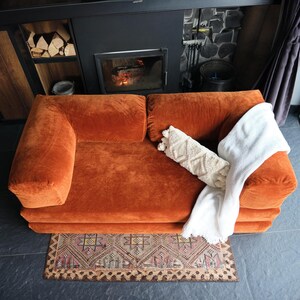 K&ouml;nnte beinhalten: Ein orangefarbenes Cordsofa mit zwei Kissen, einer wei&szlig;en Decke und einem cremefarbenen Zierkissen. Ein gemusterter Teppich liegt vor dem Sofa. Ein Kamin und Brennholz sind im Hintergrund zu sehen.