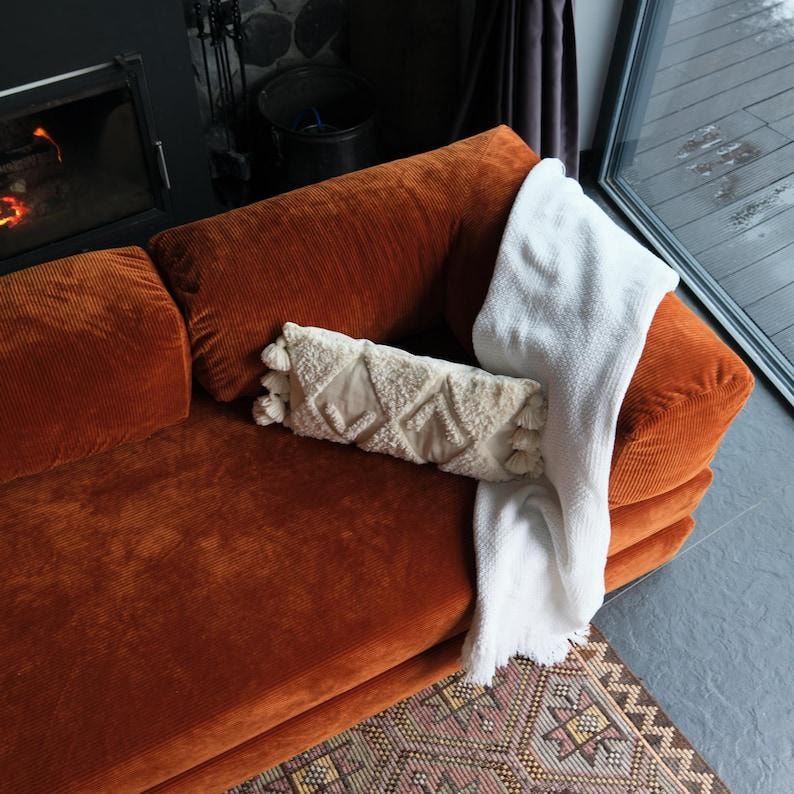 K&ouml;nnte beinhalten: Ein orangefarbenes Cordsofa mit einer cremefarbenen Decke und einem dekorativen Kissen. Das Kissen hat ein geometrisches Muster und Quasten. Ein gemusterter Teppich ist im Vordergrund zu sehen.