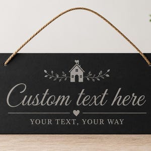 Puede incluir: Letrero rectangular negro con un diseño decorativo de casa y hojas. El letrero dice "Custom text here" y "Your text, your way" en una fuente gris claro. Se utiliza una cuerda marrón para colgarlo.
