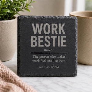 May include: A square black slate coaster with rough, natural edges. The surface features engraved text that reads: WORK BESTIE noun. The person who makes work feel less like work. see also: Sarah. The lettering is in a light grey, contrasting colour.