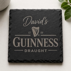 May include: Black square slate coaster with the name "David's" in cursive script above the Guinness logo. The logo includes a harp and the words "GUINNESS DRAUGHT". The coaster has a rough, textured surface.