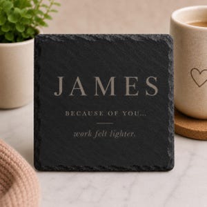May include: A square black slate coaster with rough, textured edges. The text reads: JAMES, BECAUSE OF YOU..., work felt lighter. The lettering is in a light grey, serif font, centred on the surface of the dark stone material.