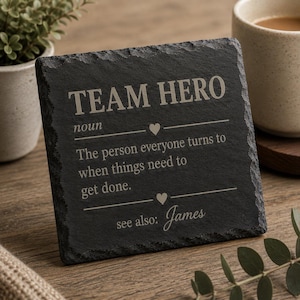 Pode incluir: Um porta-copos quadrado de ardósia preta com texto gravado que diz TEAM HERO noun, The person everyone turns to when things need to get done, see also: James, com pequenos ícones de coração separando as linhas de texto.
