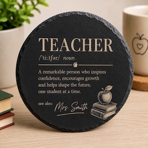 May include: A round black slate coaster featuring the engraved word TEACHER, its phonetic spelling, and a definition describing a remarkable person who inspires confidence. It includes a graphic of an apple on two books and the text see also: Mrs Smith.