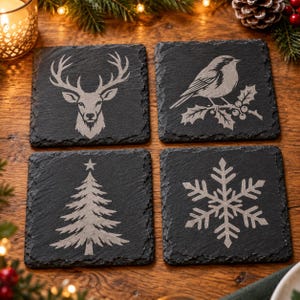 May include: A set of four square black slate coasters with rough, natural edges. Each coaster features a light grey engraved design: a deer head, a bird perched on a holly branch, a Christmas tree topped with a star, and a detailed snowflake.