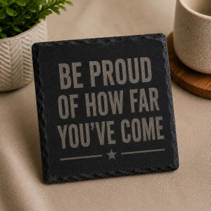 May include: A square black slate coaster with the text "BE PROUD OF HOW FAR YOU'VE COME" in grey lettering. A small star and a line are below the text. The coaster has a rough, textured edge.