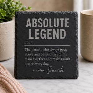 Könnte beinhalten: Ein quadratischer Untersetzer aus schwarzem Schiefer mit rauen, strukturierten Kanten. Die Oberfläche zeigt eingravierten weißen Text: ABSOLUTE LEGEND noun The person who always goes above and beyond, keeps the team together and makes work better every day. see also: Sarah