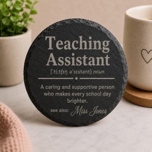 May include: A circular black slate coaster with the words "Teaching Assistant" in white. The definition of a teaching assistant is included, along with the name "Miss Jones". A mug and plant are in the background.