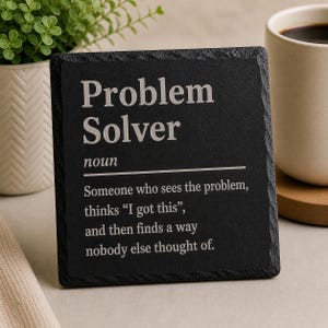 Puede incluir: Un posavasos cuadrado negro con las palabras "Problem Solver" en blanco y una definición. Una taza de café y una pequeña planta en maceta están al lado del posavasos.