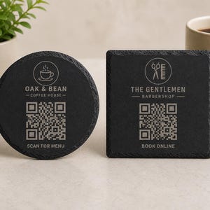 May include: Two black slate coasters, one round and one square, with etched text and QR codes. The round coaster reads "OAK & BEAN - COFFEE HOUSE - SCAN FOR MENU." The square coaster reads "THE GENTLEMEN - BARBERSHOP - BOOK ONLINE."