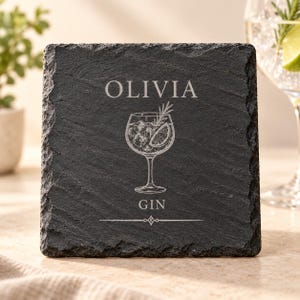 May include: A square black slate coaster with rough, natural edges. The center features an engraved illustration of a cocktail glass with ice and a lime slice, the name OLIVIA, the word GIN, and a decorative horizontal line below.