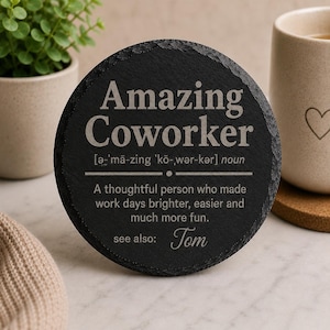 Puede incluir: Un posavasos redondo de pizarra negra con la inscripción "Amazing Coworker" en gris claro. El posavasos incluye una definición y el nombre "Tom". Una taza y una planta en maceta están en el fondo.