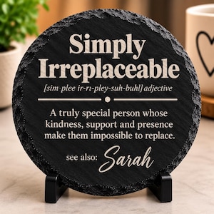 Pode incluir: Uma placa redonda de ardósia preta com uma borda rugosa e texturizada, exibida num suporte preto. A superfície apresenta texto gravado a branco que define a palavra Simply Irreplaceable, uma frase descritiva e o nome Sarah em letra cursiva.