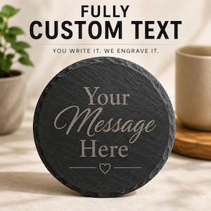 May include: A round black slate coaster with a rough, textured edge. The centre is engraved with the text "Your Message Here" in a script and serif font combination, featuring a small heart icon centred below the text on a horizontal line.