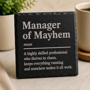 Könnte beinhalten: Ein schwarzer quadratischer Untersetzer mit den weißen Worten "Manager of Mayhem" und einer Definition. Der Untersetzer besteht aus einem dunklen, strukturierten Material und wird neben einer Kaffeetasse und einer kleinen Topfpflanze gezeigt.