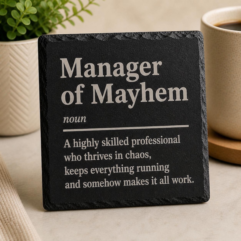 May include: A black square coaster with the words "Manager of Mayhem" in white, along with a definition. The coaster is made of a dark, textured material and is displayed next to a coffee mug and a small potted plant.
