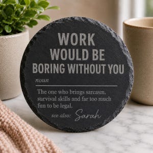 May include: A round black slate coaster with rough, textured edges. The surface features engraved white text that reads: WORK WOULD BE BORING WITHOUT YOU, noun, The one who brings sarcasm, survival skills and far too much fun to be legal, see also: Sarah.