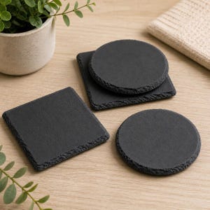 May include: A collection of black slate coasters in various shapes: square and round. The coasters have a rough, textured edge and sit on a light wood surface. A small potted plant and a folded beige cloth are in the background.
