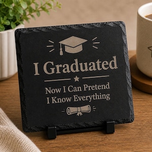 Puede incluir: Un posavasos cuadrado de pizarra negra con el texto "I Graduated Now I Can Pretend I Know Everything" en una fuente gris claro. El posavasos presenta un gorro de graduación y un gráfico de diploma. El posavasos se muestra en un soporte negro.