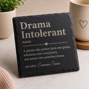May include: A square black slate coaster with rough, chiseled edges. The surface features white engraved text that reads: Drama Intolerant noun, a person who prefers facts over gossip, solutions over complaints, and peace over pointless drama, see also: Common Sense.