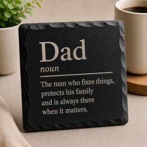Può includere: Un sottobicchiere in ardesia nera con bordo decorativo. La parola "Dad" è incisa in grassetto, seguita dalla definizione: "L'uomo che aggiusta le cose, protegge la sua famiglia ed è sempre presente quando conta."