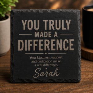 May include: A square black slate coaster with rough, natural edges. The text is engraved in a light tan color and reads: YOU TRULY MADE A DIFFERENCE, Your kindness, support and dedication make a real difference. Sarah. The surface is matte and dark.