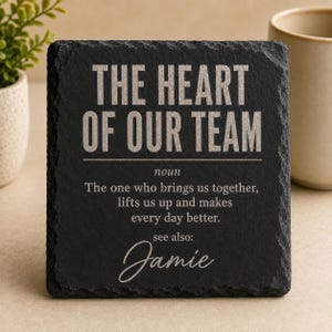 May include: A square black slate coaster with rough, chiseled edges. The text reads: THE HEART OF OUR TEAM, noun, The one who brings us together, lifts us up and makes every day better. see also: Jamie. The lettering is engraved in a light grey colour.