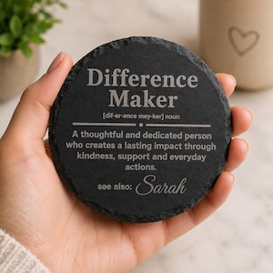 以下が含まれることがあります： 「Difference Maker」の文字が白で刻まれた、丸い黒いスレートコースター。コースターには定義と「Sarah」という名前が記載されています。背景にはハートのデザインのマグカップがあります。