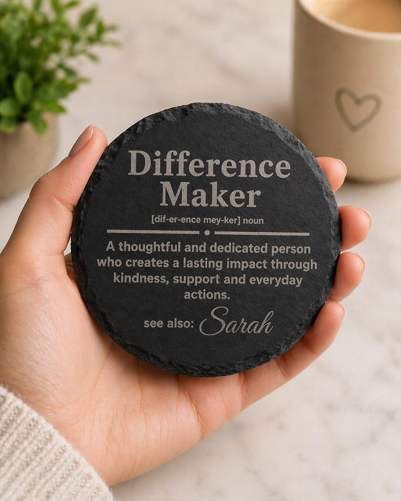 以下が含まれることがあります： 「Difference Maker」の文字が白で刻まれた、丸い黒いスレートコースター。コースターには定義と「Sarah」という名前が記載されています。背景にはハートのデザインのマグカップがあります。