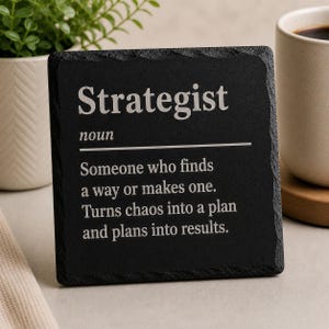 Könnte beinhalten: Schwarzer Schiefer-Untersetzer mit dem Wort "Strategist" in Weiß und seiner Definition. Daneben sind eine weiße Tasse und eine Topfpflanze zu sehen.