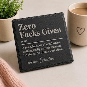 May include: A black square coaster with white text reading "Zero Fucks Given" and a definition. The word "Freedom" is also present. A beige mug with a heart design is visible.