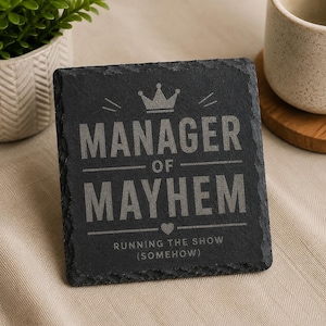 Könnte beinhalten: Ein quadratischer, schwarzer Schieferuntersetzer mit den Worten "MANAGER OF MAYHEM" in fetter, serifenloser Schrift. Eine Kronengrafik befindet sich über dem Wort "MANAGER". Darunter ist der Text "RUNNING THE SHOW (SOMEHOW)" aufgedruckt.