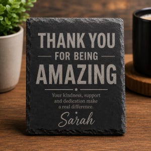 May include: A rectangular black slate plaque with rough, chiseled edges. The text reads: THANK YOU FOR BEING AMAZING. Your kindness, support and dedication make a real difference. Sarah. The engraving is in a light grey colour against the dark stone.