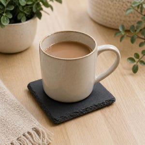 May include: A beige ceramic mug filled with a light brown liquid, resting on a square black slate coaster. The mug has a rounded shape and a handle. The coaster has a rough, textured edge. A small plant in a beige pot is in the background.