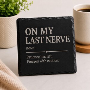 May include: A black slate coaster with the text "ON MY LAST NERVE" in white. The definition of the phrase is included below the text. A mug of coffee and a plant are in the background.