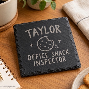 May include: A square black slate coaster with rough, uneven edges. The surface is engraved with the name Taylor, a graphic of a cookie with a bite taken out of it, and the text Office Snack Inspector in a clean, sans-serif font.