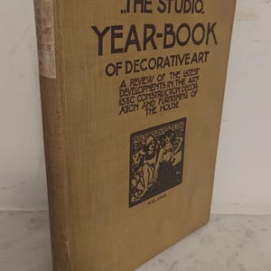 May include: A vintage book titled "THE STUDIO YEAR-BOOK OF DECORATIVE ART" with a tan cover. The cover features black text and a small illustration. The book's spine has a faded label. The book is a review of the latest developments in art.