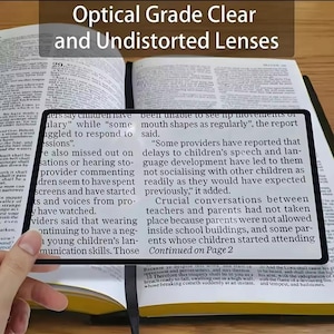 May include: A rectangular magnifying glass with a black frame is held over an open book. The text "Optical Grade Clear and Undistorted Lenses" is at the top. The book's pages are filled with printed text.