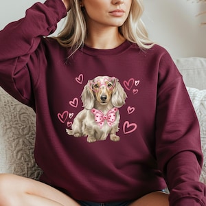 May include: Burgundy sweatshirt with a dachshund illustration wearing pink glasses and a bow tie. Pink hearts are scattered around the dog. The sweatshirt is a plain burgundy colour.