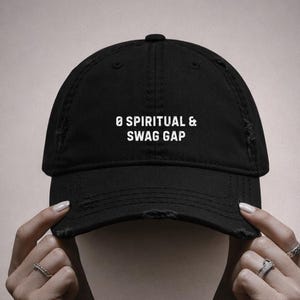 Könnte beinhalten: Schwarze Baseballkappe mit ausgefransten Rändern. Auf der Kappe steht in weißer Schrift "Ø SPIRITUAL & SWAG GAP". Die Kappe wird von zwei Händen mit Ringen gehalten.