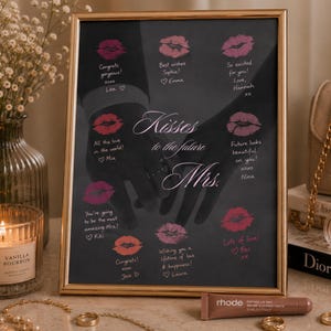 Könnte beinhalten: Ein gerahmter Druck mit goldenem Rand zeigt den Text "Kisses to the future Mrs." und Lippenstiftküsse. Handschriftliche Nachrichten umgeben den Text. Eine brennende Kerze und Schmuck sind im Vordergrund.