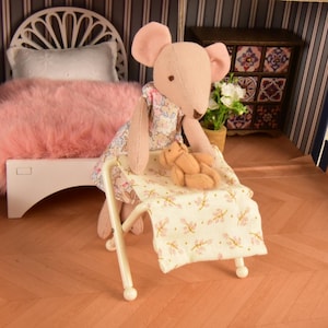 May include: A handmade dollhouse scene featuring a pink mouse doll wearing a floral dress, tending to a smaller doll on a miniature bed. The bed has a floral patterned blanket. The scene includes a white bed with a pink fluffy blanket.