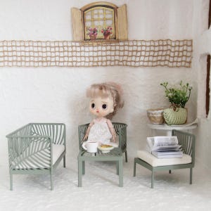 May include: A doll sits at a miniature table set with a cup and plate, surrounded by small green chairs with white cushions. The scene is set in a dollhouse with a window, flowers, and a decorative net.