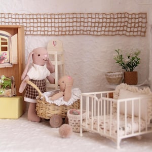 May include: A miniature nursery scene featuring a standing bunny doll in a white sweater and plaid skirt, and a bunny doll in a wicker basket. The scene includes a white crib, a small chair, and decorative plants.