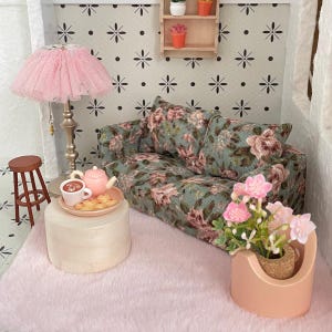 May include: Miniature dollhouse living room scene with a floral patterned sofa, pink rug, and a pink lampshade. A small table holds a teapot, cup, and pastries. A small stool and a flower pot with pink flowers complete the scene.