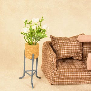 May include: A miniature scene featuring a small sofa with brown plaid cushions and a decorative plant. The plant is in a cork pot on a gray metal stand. The white flowers and green leaves add a touch of nature to the scene.