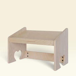 May include: A light-colored wooden stand with a flat top surface and two side panels featuring heart-shaped cutouts at the base. The front edge of the stand displays a small, engraved logo. The structure is held together by visible black screws.